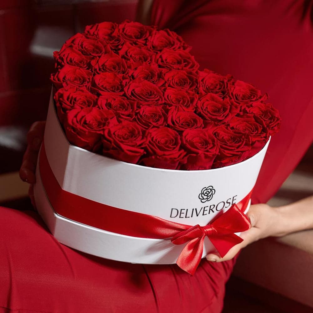 A heart-shaped white Deliverose box filled with deep red preserved roses, held in both hands