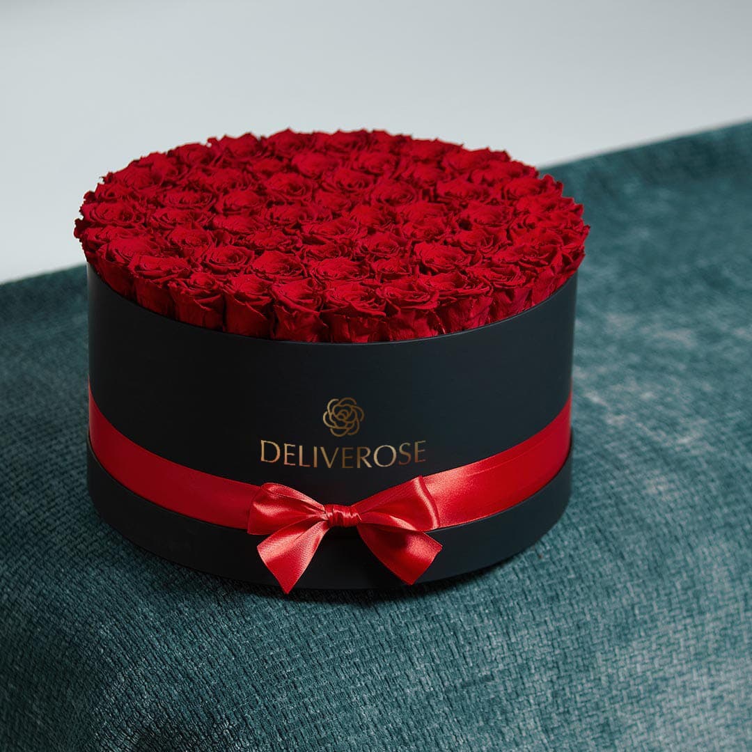 A wide black Deliverose box packed with preserved red roses, tied with a red satin ribbon and bow, on teal upholstery